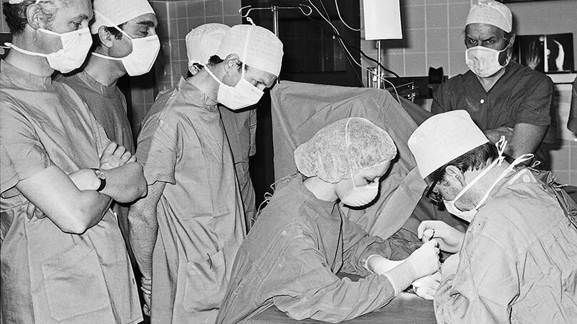 Die Schwarz-Weiß-Fotografie zeigt einen Operationssaal, in dem ein ein medizinisches Team einen nicht erkennbaren Patienten operiert. Hinter dem Operateur und der Operateurin stehen weitere Mediziner, die bei der OP zusehen.